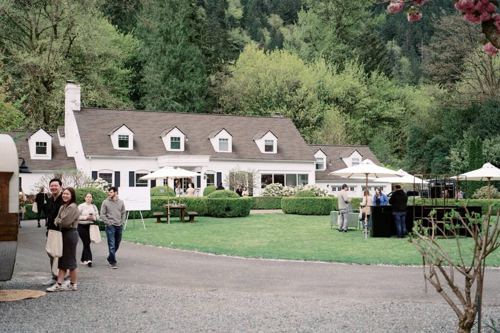 Wide view of the white farmhouse estate at Fox Hollow Farm, a luxury wedding venue, with guests on the lawn under umbrellas beside the gravel drive
