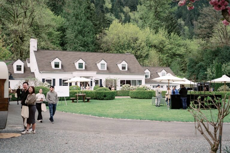Wide view of the white farmhouse estate at Fox Hollow Farm, a luxury wedding venue, with guests on the lawn under umbrellas beside the gravel drive