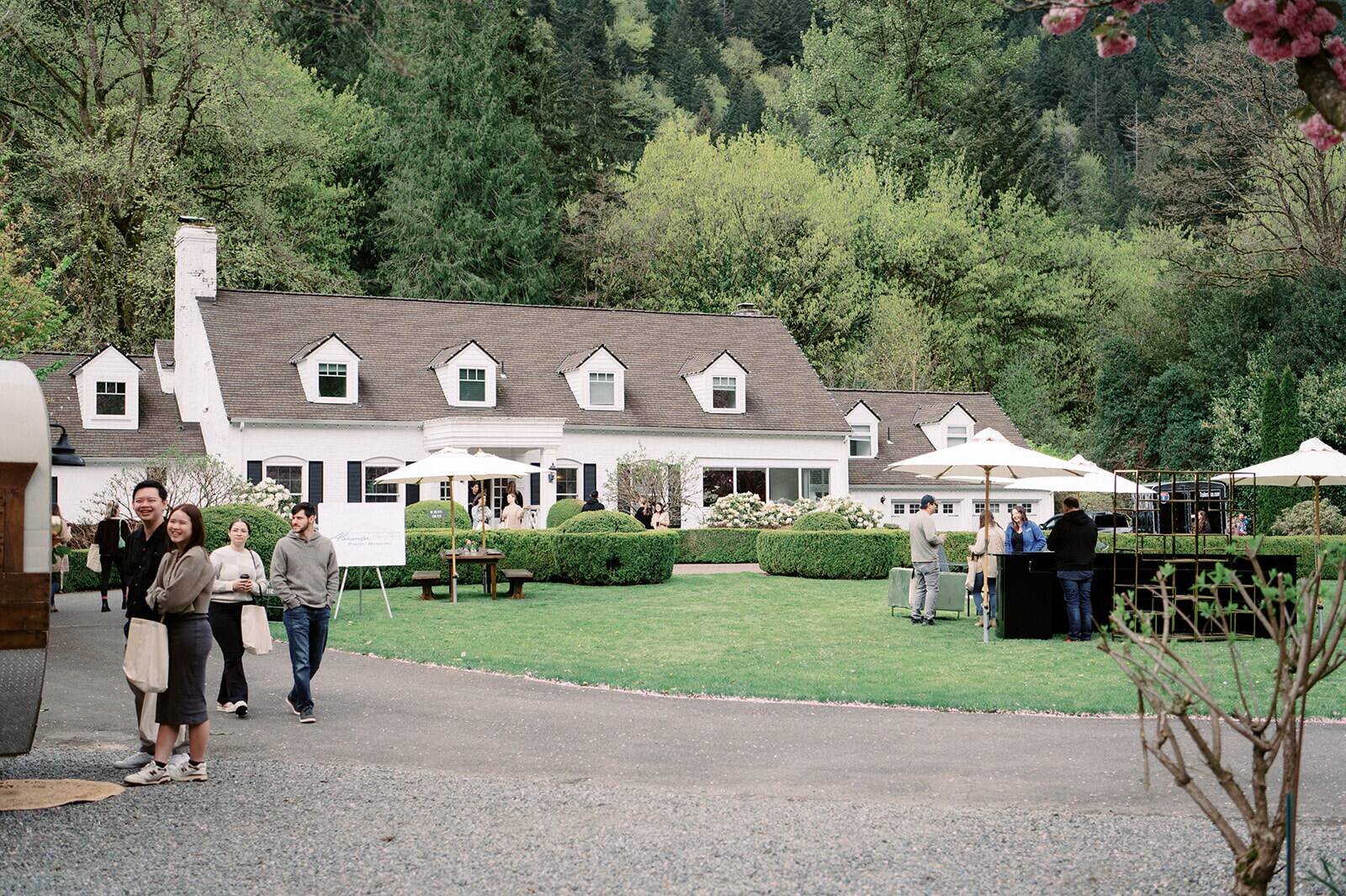 Wide view of the white farmhouse estate at Fox Hollow Farm, a luxury wedding venue, with guests on the lawn under umbrellas beside the gravel drive